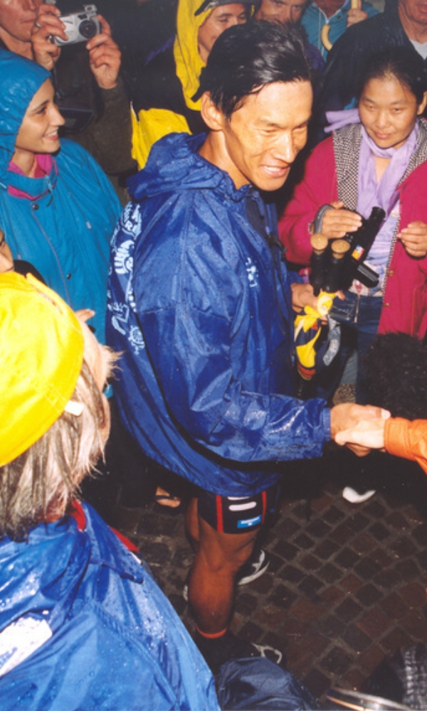 A man in a wet blue rain jacket is standing smiling and shaking someone's hand while surrounded by people in rain gear