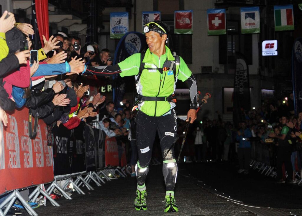 A man in a neon green rain jacket, reflective black running tights, green shoes and a headlamp runs down a dark street towards the camera while high fiving spectators at the finish line of UTMB