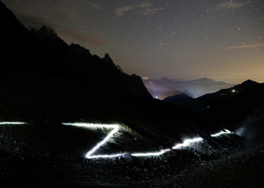 A dark mountain vista shows silhouettes of peaks while a long exposure effect of headlamps shows a lit up path of the trail through the mountains