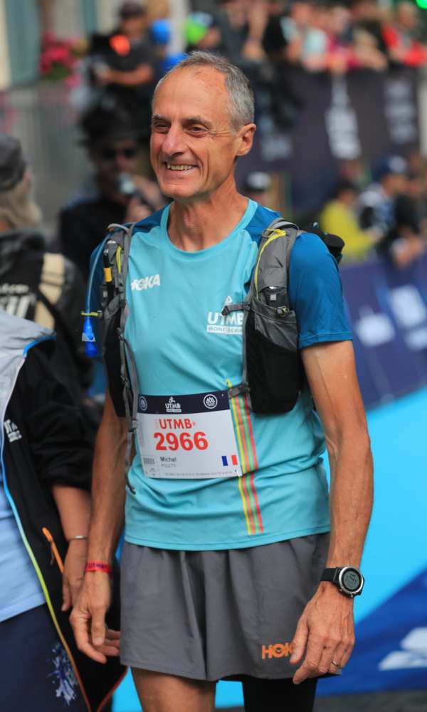 A man with gray hair wearing a blue short sleeve shirt, gray hoka shorts and a hydration vest smiles with an out of focus crowd in the background
