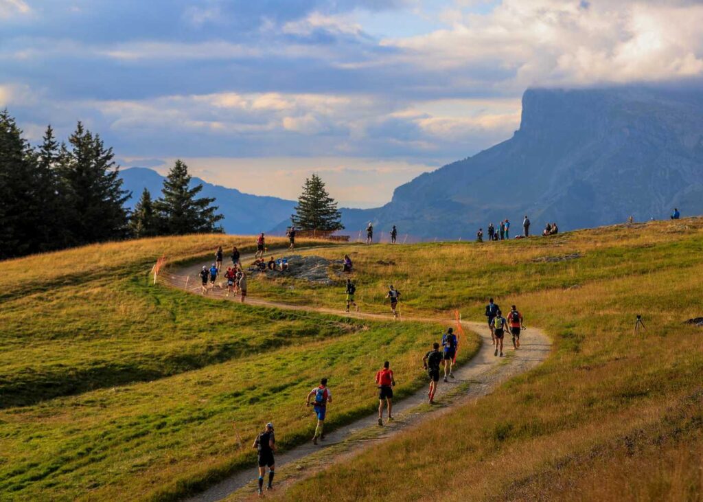 A line of runners trot up a wide grassy doubletrack slope on the UTMB course with mountains in the distance