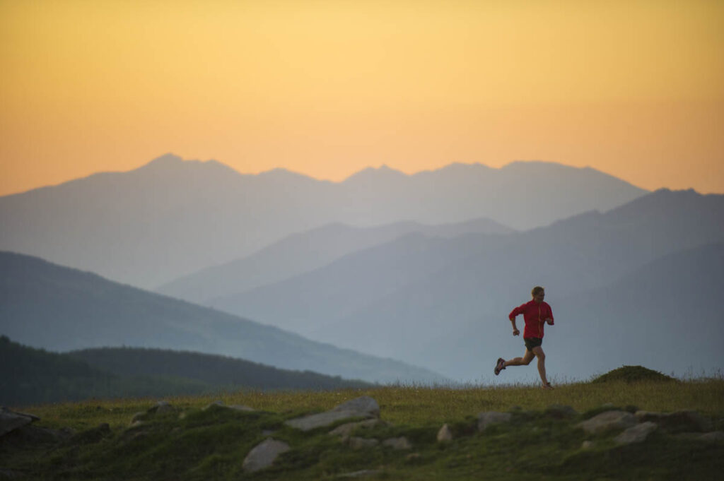 A person runs midstride across a grassy slope wearing a red jacket and running shorts. Behind them are layers of blue shadowed mountain peaks with an orange sky beyond.