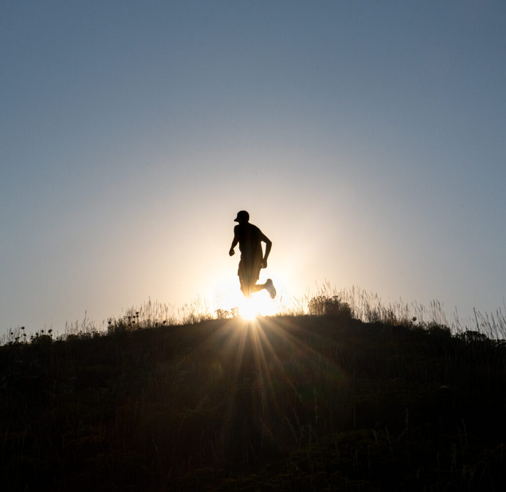 A person is running atop a hill, silhouetted by the setting sun.