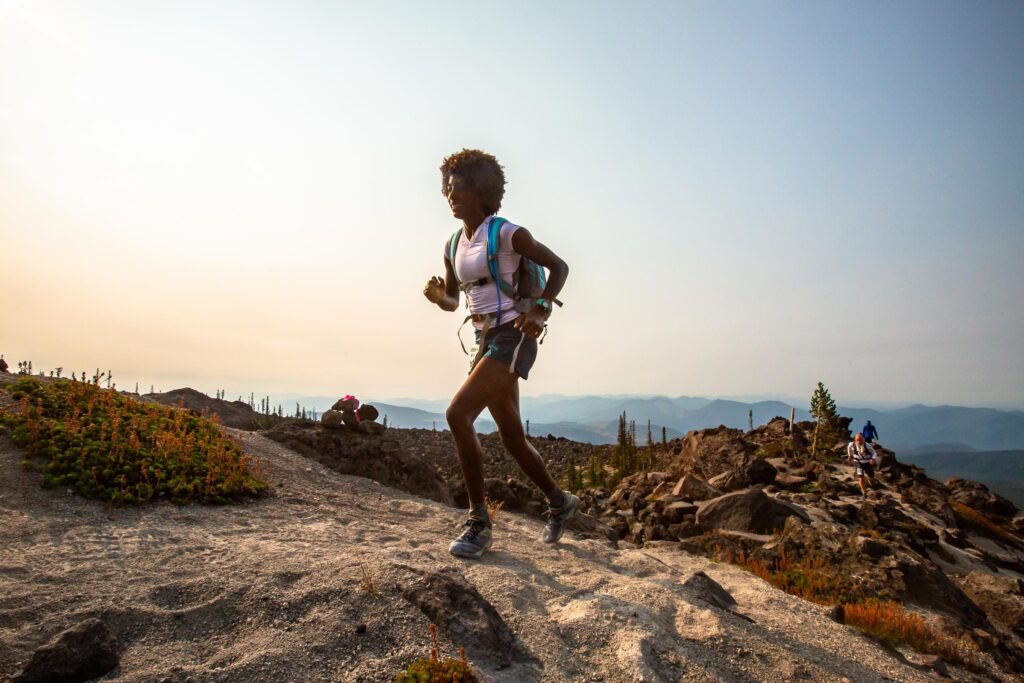A woman is running uphill over sandy rocks wearing shorts, a white short sleeve shirt and a blue backpack. In the background is a boulder covered landscape with sparse pine trees and two runners traversing a ridgeline.